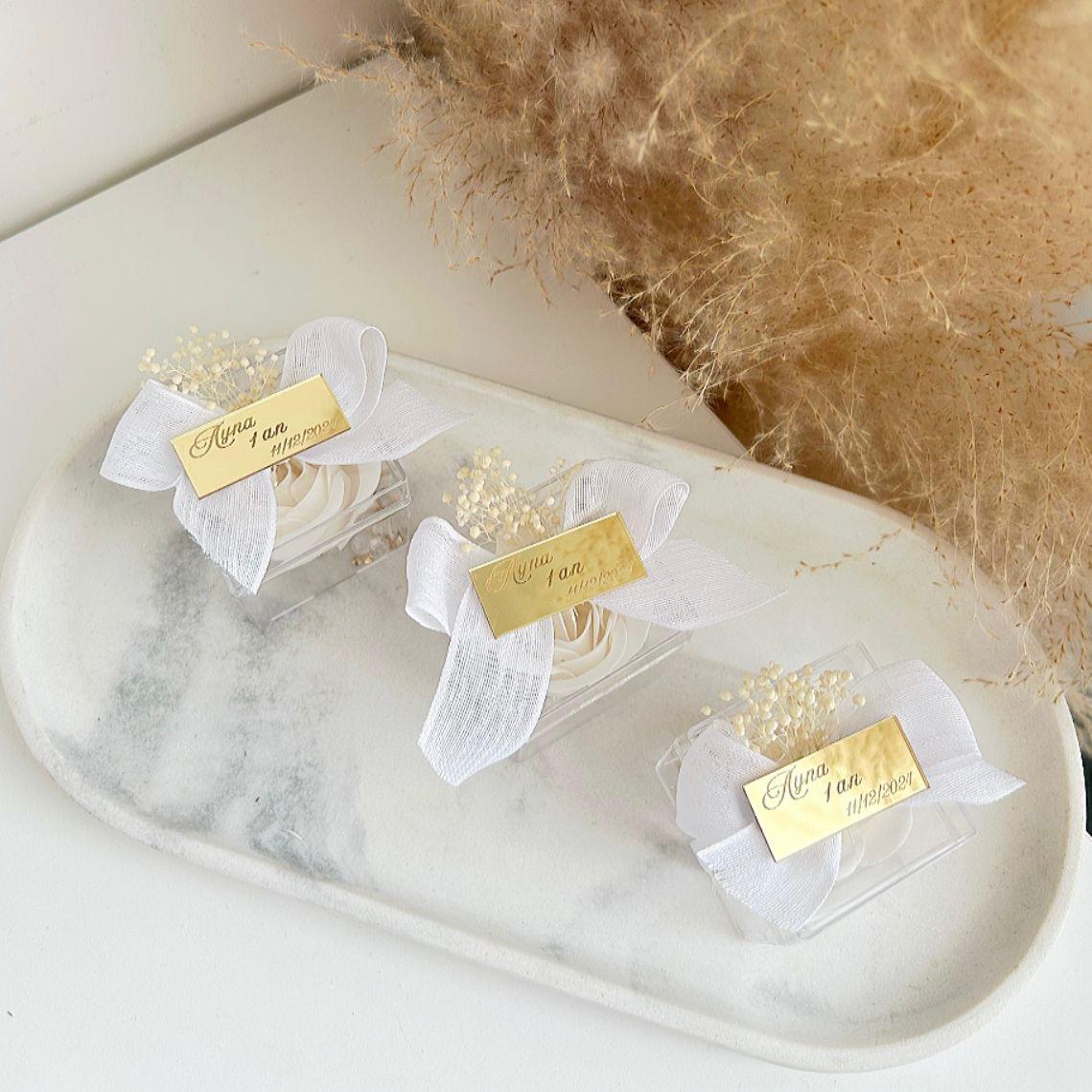 Lot de roses de savon Angelita en boîtes transparentes, décorées de gypsophile, avec nœud en ruban blanc et plaque dorée personnalisée, présentées sur un plateau.