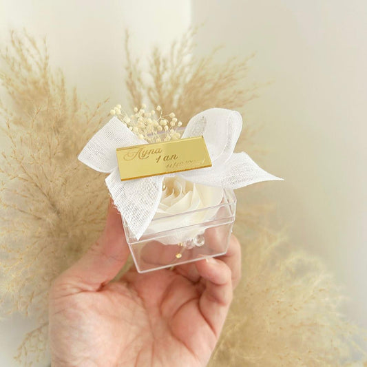 Lot de roses de savon Angelita en boîtes transparentes, décorées de gypsophile, avec nœud en ruban blanc et plaque dorée personnalisée, présentées sur un plateau.