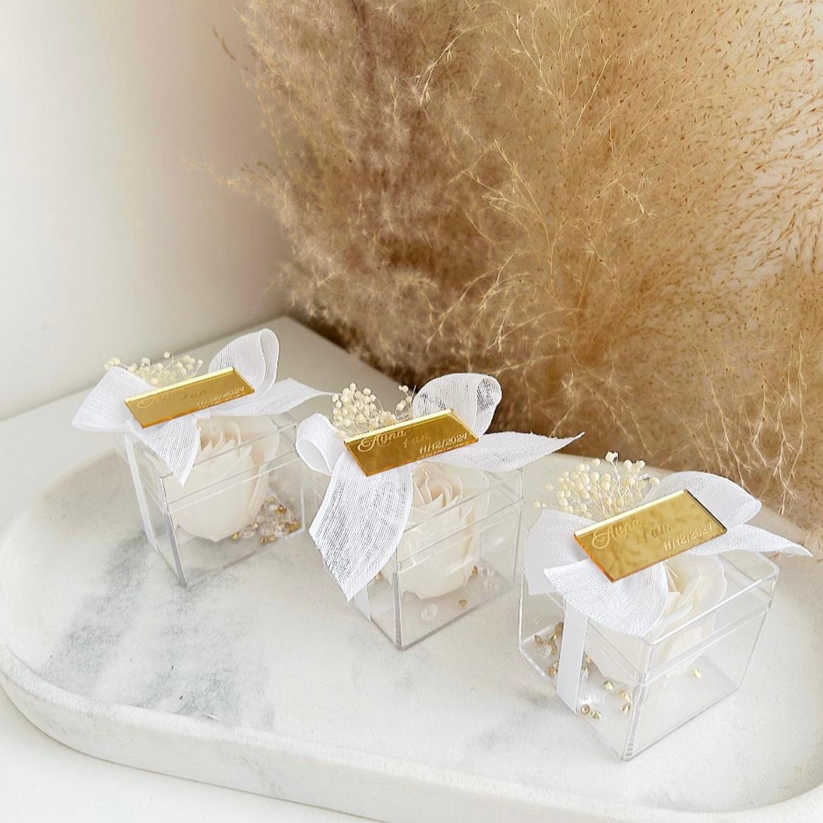 Lot de roses de savon Angelita en boîtes transparentes, décorées de gypsophile, avec nœud en ruban blanc et plaque dorée personnalisée, présentées sur un plateau.